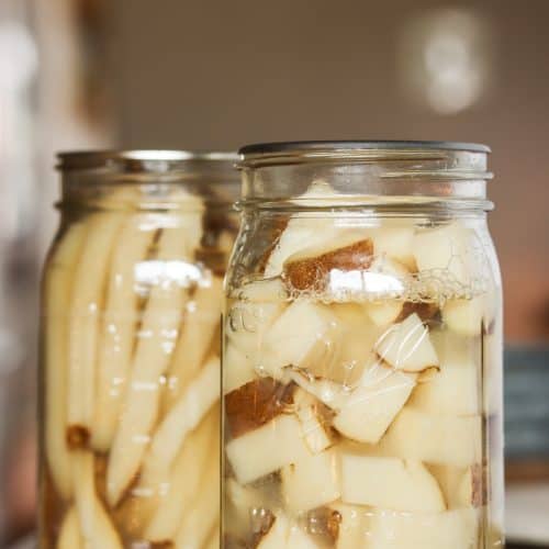 Two mason jars filled with pressure-canned potatoes with skins on, one jar containing cubed potatoes and the other with potato strips, sitting on a wooden surface with a blurred background.