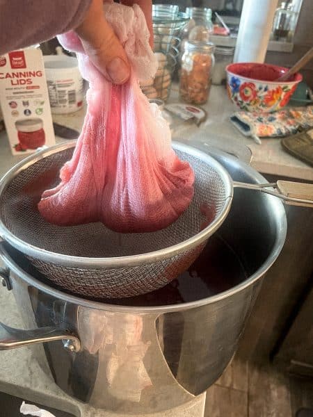 Large stainless steel pot with a fine mesh strainer on top with cheesecloth being held up to remove the pulp from the cranberry juice.