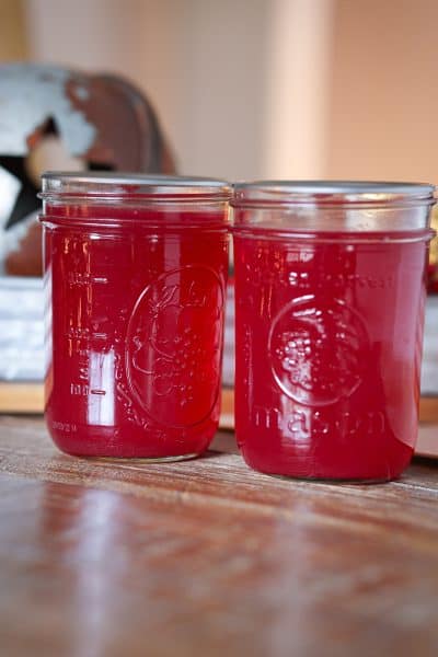 Two jars of homemade cranberry juice concentrate in a clear mason jar with a silver lid, resting on a rustic wooden surface with a softly lit background.