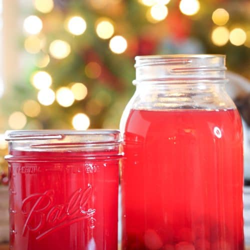 Two mason jars filled with vibrant homemade cranberry juice concentrate and cranberry juice, one with whole cranberries at the bottom, set against a warm, festive background with glowing holiday lights.